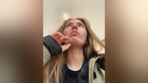 Media: Video of a young woman with long, light brown hair, wearing a black top and green jacket, looking upward with a thoughtful expression, indoors.