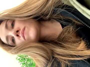 Media: Video of a young Caucasian woman with long, blonde hair and fair skin, wearing a black jacket, captured from a low angle, with blurred green foliage in the background.