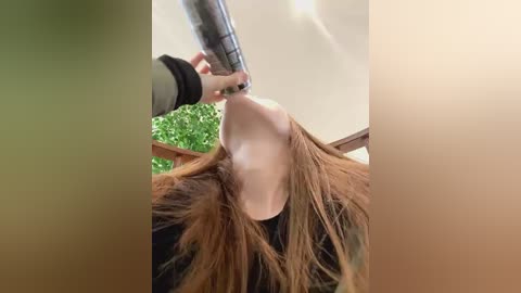 Media: Video of a young woman with long, straight brown hair, lying on her back with her head tilted back, receiving a drink from a metal straw held by an unseen person. Background shows a ceiling and green foliage.