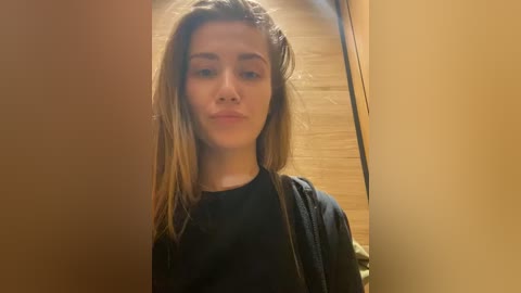 Media: Video of a young Caucasian woman with long, straight brown hair, wearing a black top and black jacket, standing indoors with wooden walls and a mirror. She makes a pouty face.