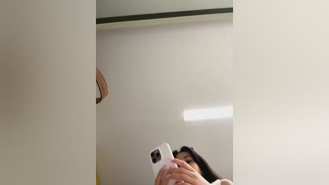 Media: A video shows a woman with long black hair taking a selfie in a minimalist, white-walled room. She holds a white iPhone, capturing her reflection in a mirror. The room has a modern, clean aesthetic.