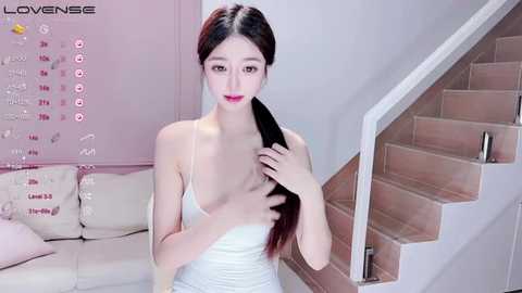 Media: Video of an Asian woman with long black hair, wearing a white tank top, posing in a modern, minimalist living room with beige furniture and a wooden staircase.