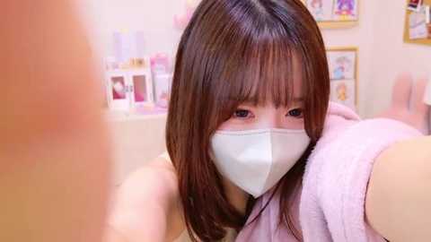 Media: Video of an Asian woman with shoulder-length brown hair and a white mask, wearing a pink robe, taking a selfie in a well-lit, pastel-colored room with framed photos and shelves.