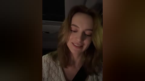 Media: Video of a smiling woman with shoulder-length, wavy blonde hair, wearing a white, patterned blouse, seated in a dimly-lit car interior.