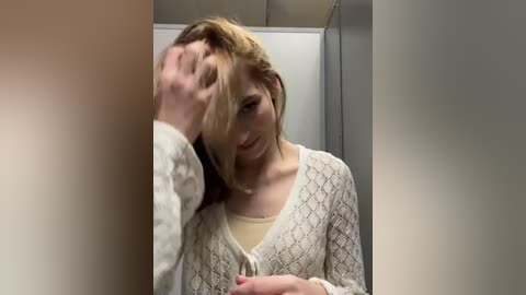 Media: Video of a pale-skinned woman with shoulder-length brown hair, wearing a light sweater, holding her head in a bathroom with tiled walls and a white sink.