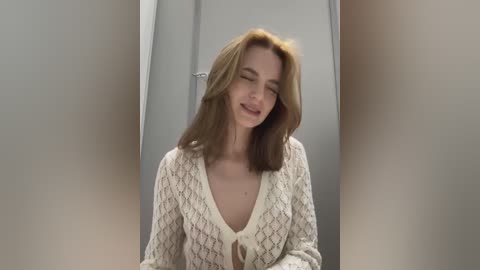 Media: Video of a young Caucasian woman with shoulder-length auburn hair, wearing a white, patterned cardigan, smiling playfully, standing in a dimly lit, narrow hallway.