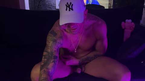 Media: Video of a shirtless tattooed man with a white baseball cap, seated on a dark couch in dim lighting. He holds a black dog, with purple and blue lights illuminating the scene.