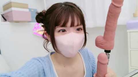 Media: Video of an Asian woman with a pale complexion, wearing a white mask, and blue sweater, holding a realistic, uncircumcised, erect penis in a clinical setting.