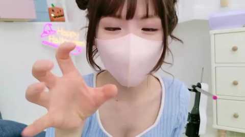 Media: Video of a young Asian woman in a light blue striped top, wearing a pink face mask, holding a glowing stick, surrounded by pastel-colored furniture.