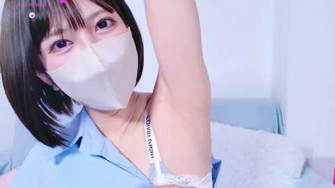 Media: Video of an Asian woman with short black hair, wearing a white surgical mask, revealing a white lace bra strap. She's in a light blue shirt, against a white background.