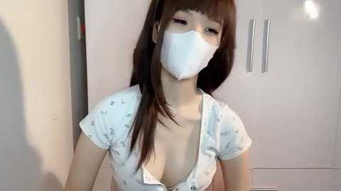 Media: Video of an East Asian woman with long brown hair, wearing a white surgical mask, and a low-cut, white floral-patterned blouse revealing ample cleavage, standing in a modern, minimalist bathroom with white cabinets and silver handles.