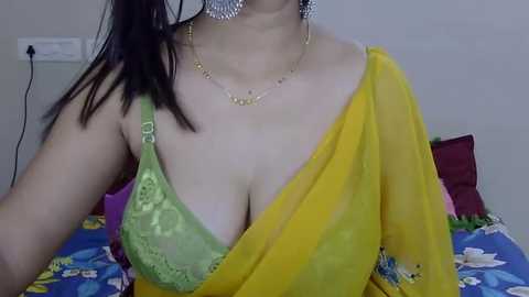 Media: Video of a woman with medium skin tone, dark hair, wearing a yellow sari with green lace bra, silver earrings, and a gold necklace. She sits on a bed with colorful pillows.