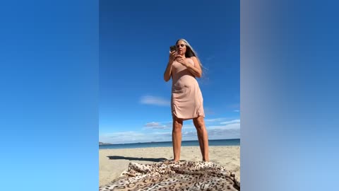 Media: Video of a middle-aged woman with long gray hair, wearing a pink dress, standing on a sandy beach under a clear blue sky.