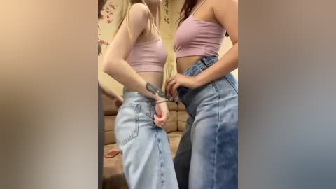 Media: Video of two young women, fair-skinned, in light denim jeans and pastel tops, embracing in a cozy living room with floral wallpaper and a brown couch.