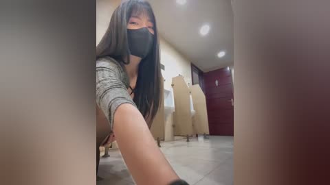 Media: Video of an Asian woman in a gray sweater and black face mask, crouching in a dimly lit room with beige walls, a wooden door, and cardboard boxes.