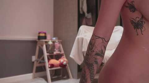 Media: Video of a nude woman with extensive tattoos, standing in a dimly lit, modern bathroom with a wooden step ladder, colorful candles, and a white towel.