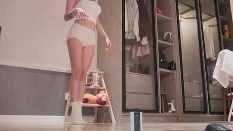 Media: Video of a woman in white lingerie, standing on a wooden stool in a bedroom with a walk-in closet and a stuffed toy on a shelf.