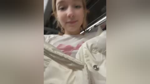 Media: Video of a young woman with light skin and light brown hair, wearing a white jacket, seated in a car. The image is blurry and out of focus.