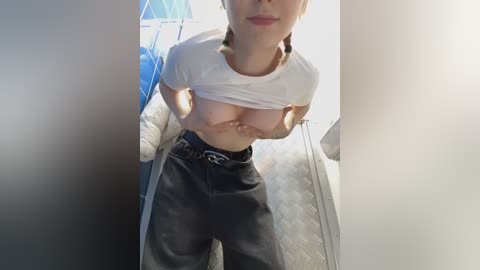 Media: A video of a fair-skinned woman lifting her white t-shirt to expose her medium-sized breasts. She wears black pants and is in a laundry room with blue and white appliances in the background.