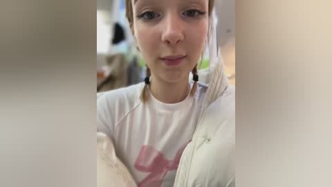 Media: Video of a young Caucasian woman with fair skin, light brown hair in pigtails, wearing a white T-shirt with a pink graphic, and a white jacket. Background is blurred, showing a kitchen with greenery.