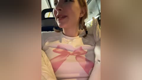 Media: A video of a young woman with light skin, wearing a white shirt with a pink bow, smiling inside a car.