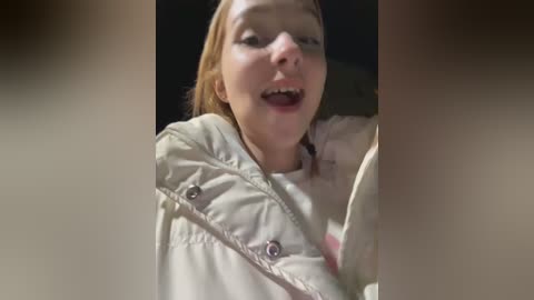 Media: Video of a young, fair-skinned girl with light brown hair, wearing a white jacket, smiling broadly while sitting in a dark car.