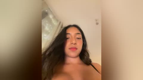 Media: Video of a young Latina woman with long, dark hair, light skin, and full lips. She's topless, wearing a black strap, in a dimly lit room with beige walls and a shiny, reflective surface in the background.