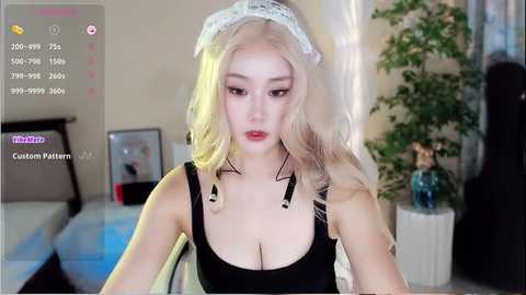 Media: Video of a fair-skinned, blonde woman with long hair, wearing a revealing black maid outfit with lace trim, in a dimly-lit room with plants and a TV.