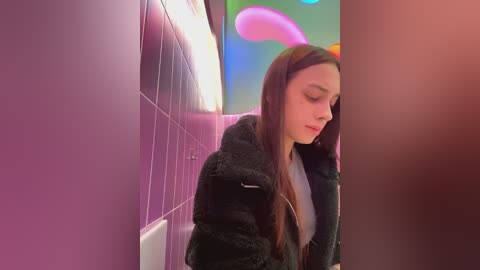 Media: A video of a young woman with long brown hair, wearing a black fur coat, standing in a dimly lit, narrow hallway with purple-tiled walls and a glowing, abstract, colorful ceiling.