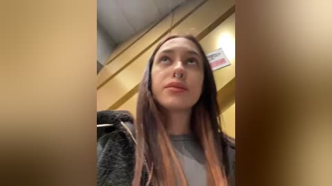 Media: A video of a young woman with long, straight, dark brown hair, light skin, and a nose ring, wearing a gray shirt and a dark jacket. She looks contemplative, with a hint of sadness in a beige, institutional setting.