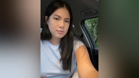 Media: Video of a young woman with long black hair, wearing a white T-shirt, sitting in a car. Background includes a blurry, green-lit window.