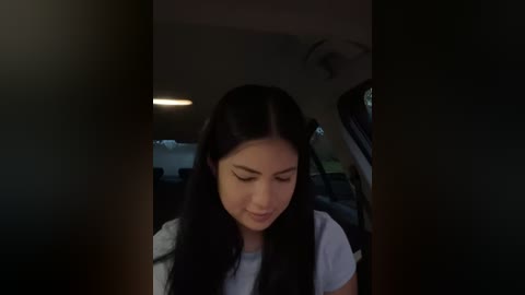 Media: Video of a young Asian woman with long black hair, wearing a white T-shirt, seated in a car with dim interior lighting, looking down.