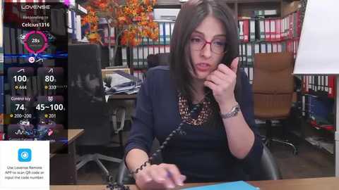 Media: Video of a middle-aged woman with glasses, dark hair, and a black top, using a phone in an office with bookshelves, desk, and a tree backdrop.