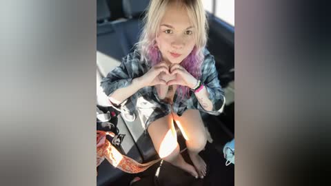 Media: A video of a blonde woman with pink hair in a car, making heart shapes with her hands, wearing a denim jacket and knee-high socks.