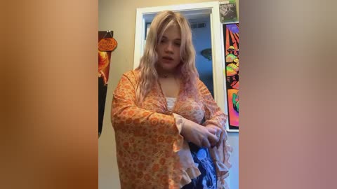 Media: Video of a plus-sized woman with long blonde hair, wearing an orange-patterned blouse and blue jeans, standing in a room with colorful wall art and a hanging orange circle.