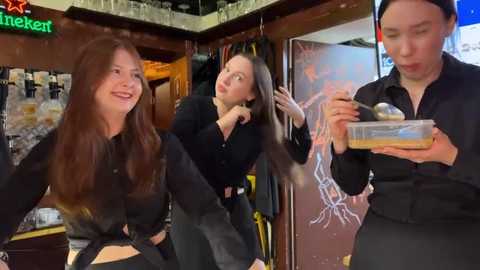 Media: Video of three women: one with long brown hair, two with short hair, in a bar. The woman with long hair is laughing, one with short hair is styling her hair, and the other is holding a bowl.