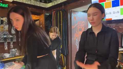 Media: Video of three women in a bar. One woman with long brown hair in black dress, another with glasses in black shirt, and a third with short hair in black shirt, all standing near a glass bar.