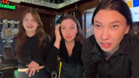 Media: Video of three young women with light skin and long hair, wearing black outfits, smiling in a cozy, dimly lit bar.