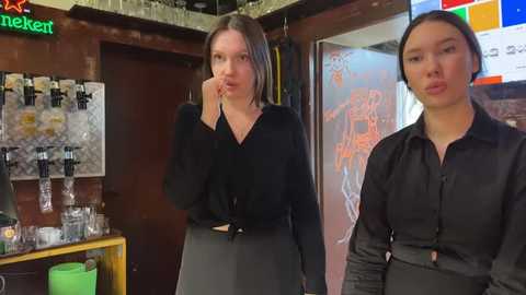 Media: Video of two women in a bar. One with long brown hair, wearing a black top and gray skirt, touches her chin. The other, with short black hair, wears a black shirt. Background includes beer taps, colorful lights, and a neon sign.