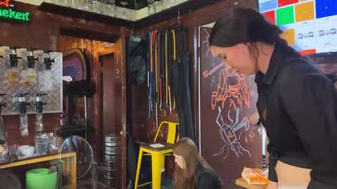 Media: Video of a woman in a black hoodie pouring beer into a glass at a bar. Background includes a wall with a chalkboard menu, hanging beers, and a colorful mosaic.