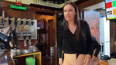 Media: A video of a young woman with long brown hair, wearing a black top and beige pants, standing in a cozy bar with beer taps, glassware, and posters in the background.