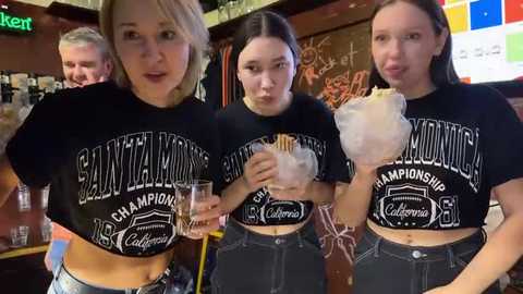 Media: Video of four women, two holding pints, in black \"Champions\" shirts, at a bar with a chalkboard and colorful flags in the background.