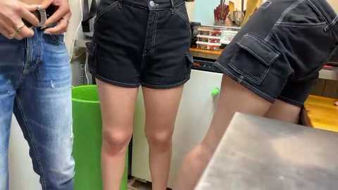 Media: Video of two women in black denim shorts and a man in blue jeans, standing in a store. The background includes a green trash can, a counter, and a guitar.