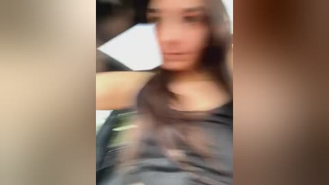 Media: A blurry, out-of-focus video of a young woman with long, dark hair, wearing a sleeveless dark top, and holding a white rectangular object. The background is indistinct.