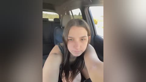 Media: A video of a young woman with long brown hair, wearing a black tank top, taking a selfie from the driver's seat of a car. The background shows a blurred view of a sunny day outside.