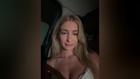 Media: Video of a blonde woman with long hair, wearing a white lace bra, inside a dimly lit car, looking slightly to the side with a neutral expression.