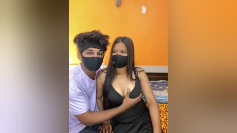 Media: Video of a young South Asian couple wearing black face masks, sitting on a bed with orange walls. The man has curly hair, a white T-shirt, and touches the woman's breast. She has long black hair, a black dress, and a shocked expression.