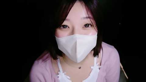 Media: Video of an East Asian woman with light skin, straight black hair, and dark eyes, wearing a white surgical mask and a pink cardigan, against a dark background.