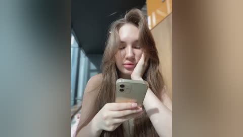 Media: Video of a young Caucasian woman with long, wavy brown hair, light skin, and full lips, wearing a white top, holding a smartphone to take a selfie, in a modern, well-lit hallway.