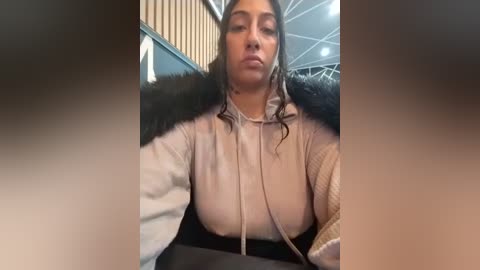 Media: Video of a woman with medium brown skin and dark hair, wearing a beige hoodie with fur trim, sitting in a modern room with beige and gray decor.